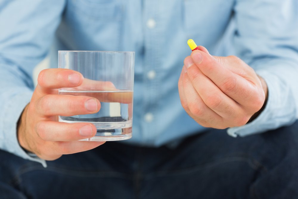 Close up of man holding a pill and glass of water in bright living room-1